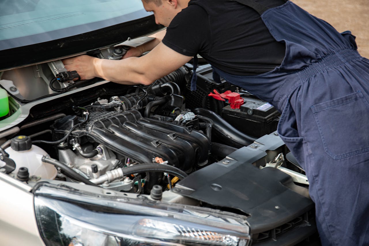 Mechanic in blue overalls working on car engine with open hood outdoors during the day.